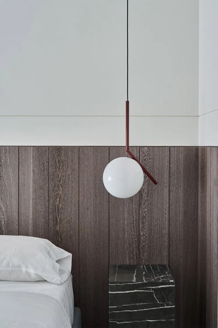 A modern minimalist bedroom featuring a single bed dressed in white bedding, a dark wooden headboard, and a pendant lamp with
