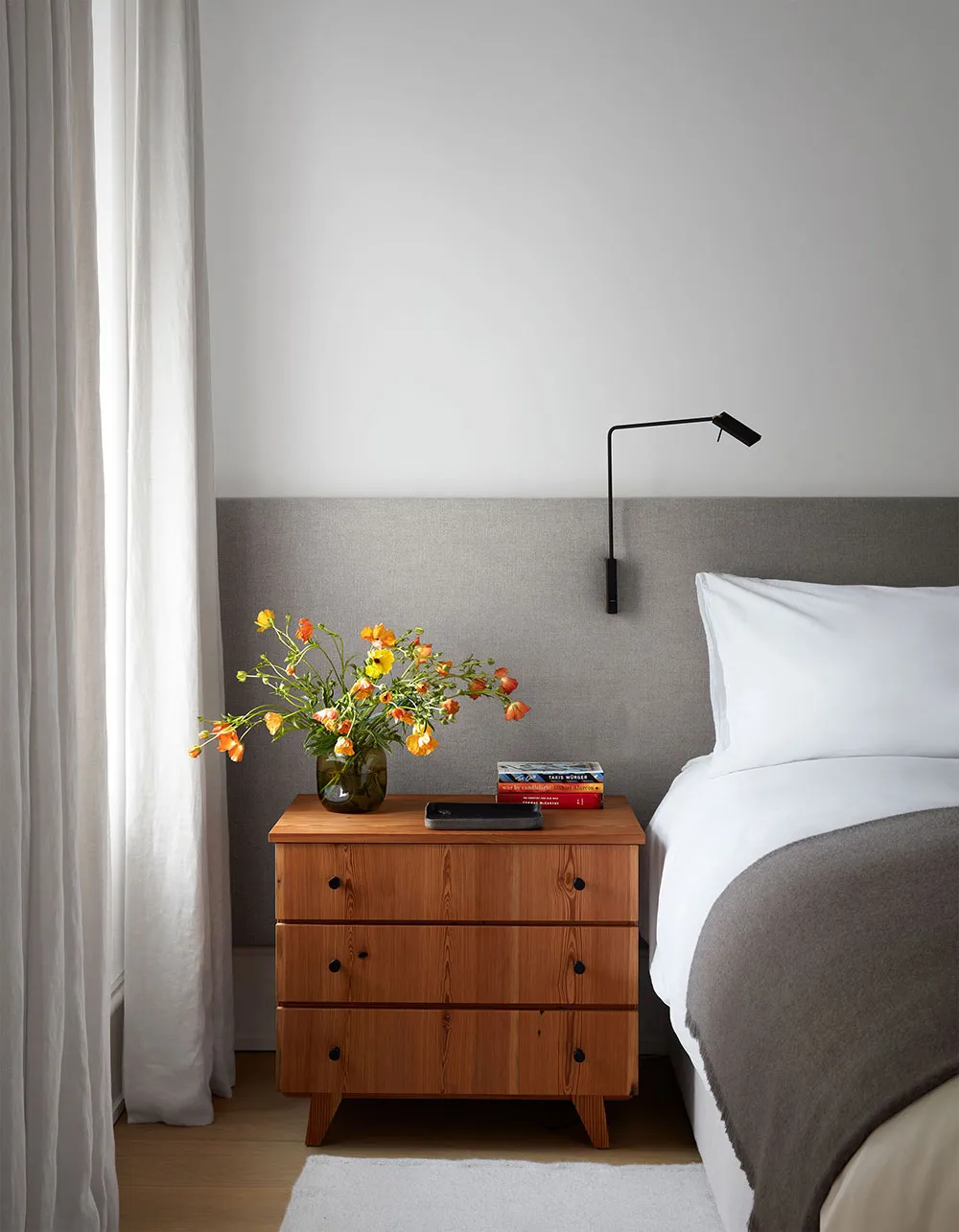 A modern bedroom featuring a wooden nightstand with a vase of flowers, books, and a small lamp.