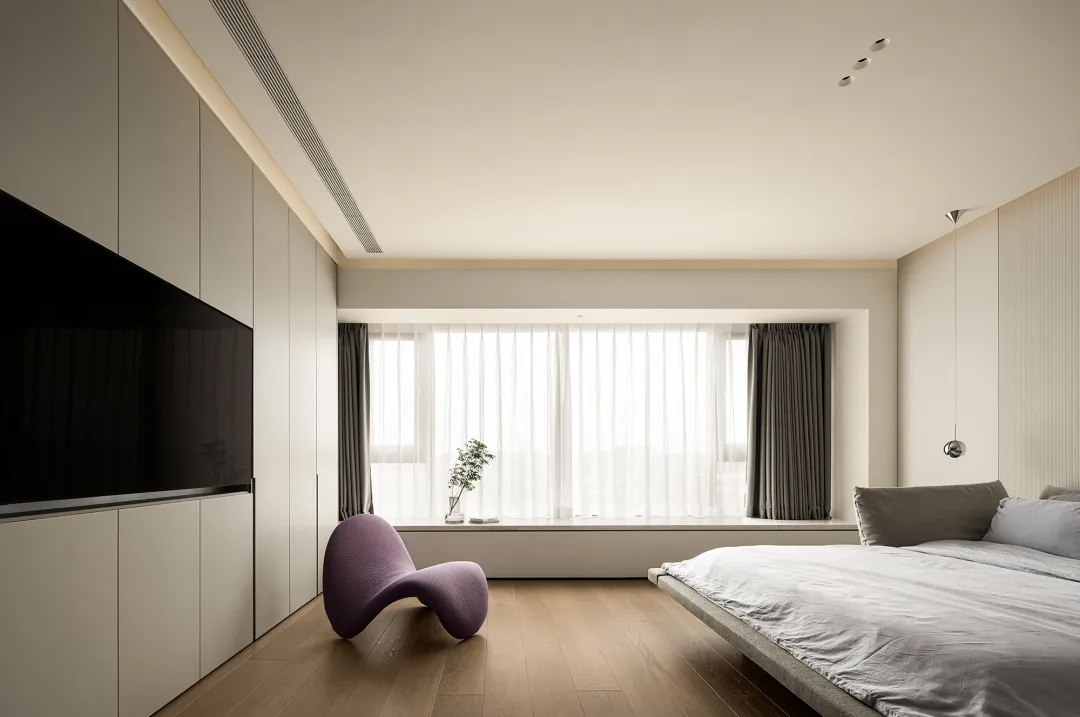 A minimalistic bedroom with light wood flooring, a modern light purple armchair, and a large window with sheer curtains.