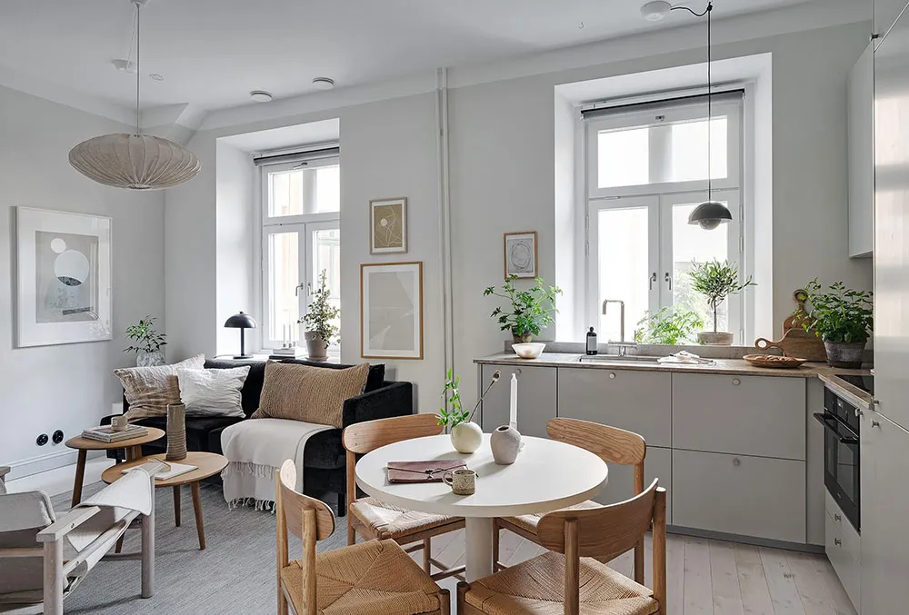 A minimalist residential space featuring a white dining table surrounded by wooden chairs, a grey sofa with white cushions, a