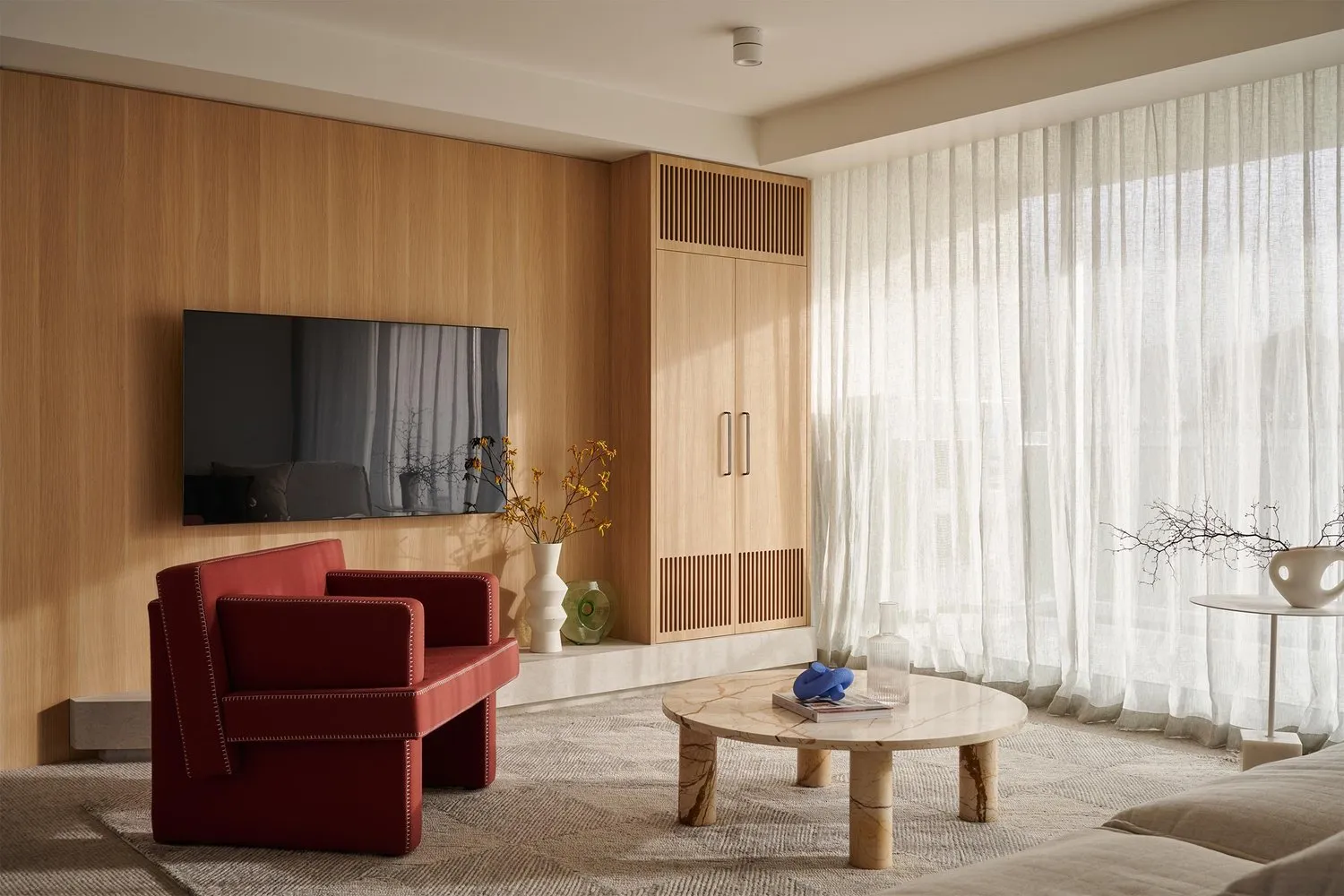 A modern living room featuring wooden walls, red armchairs, a marble coffee table, and white sheer curtains.