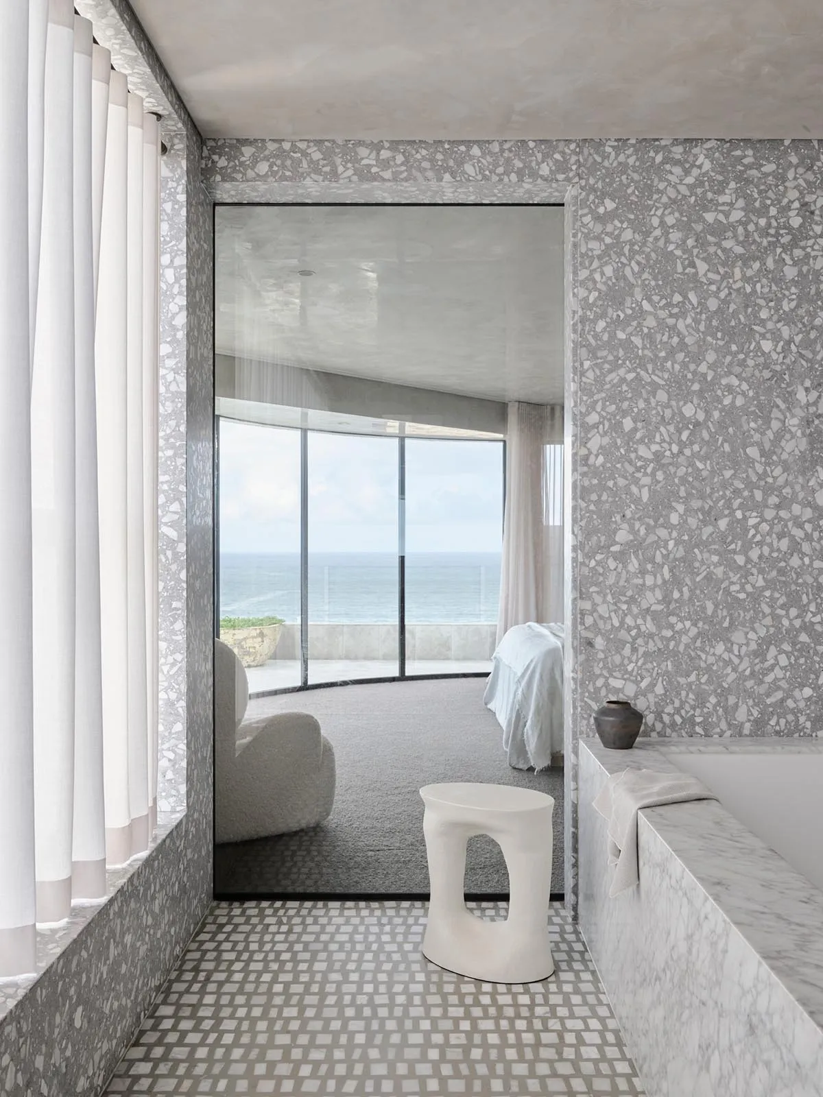 Modern minimalist bedroom with marble walls and floor, glass door leading to a balcony overlooking the ocean.
