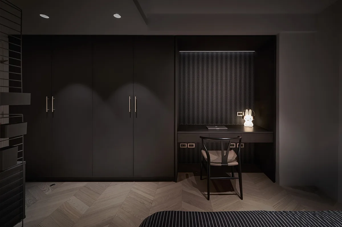 A minimalist bedroom with black furniture, wood flooring, and a desk with a light fixture.