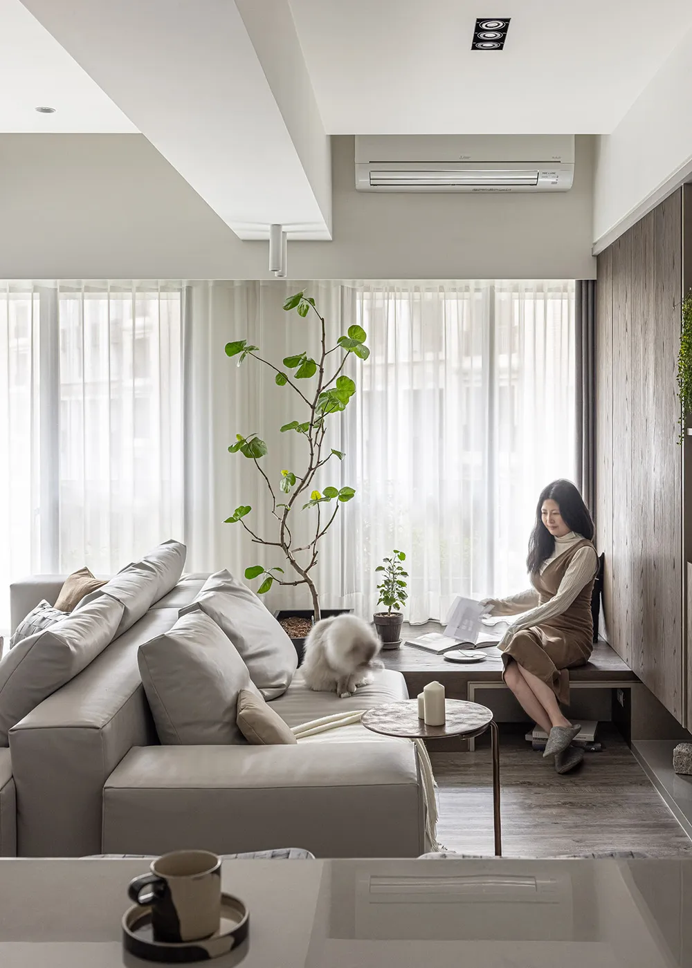 A minimalist living room with a gray sofa and a woman reading a book near a window.
