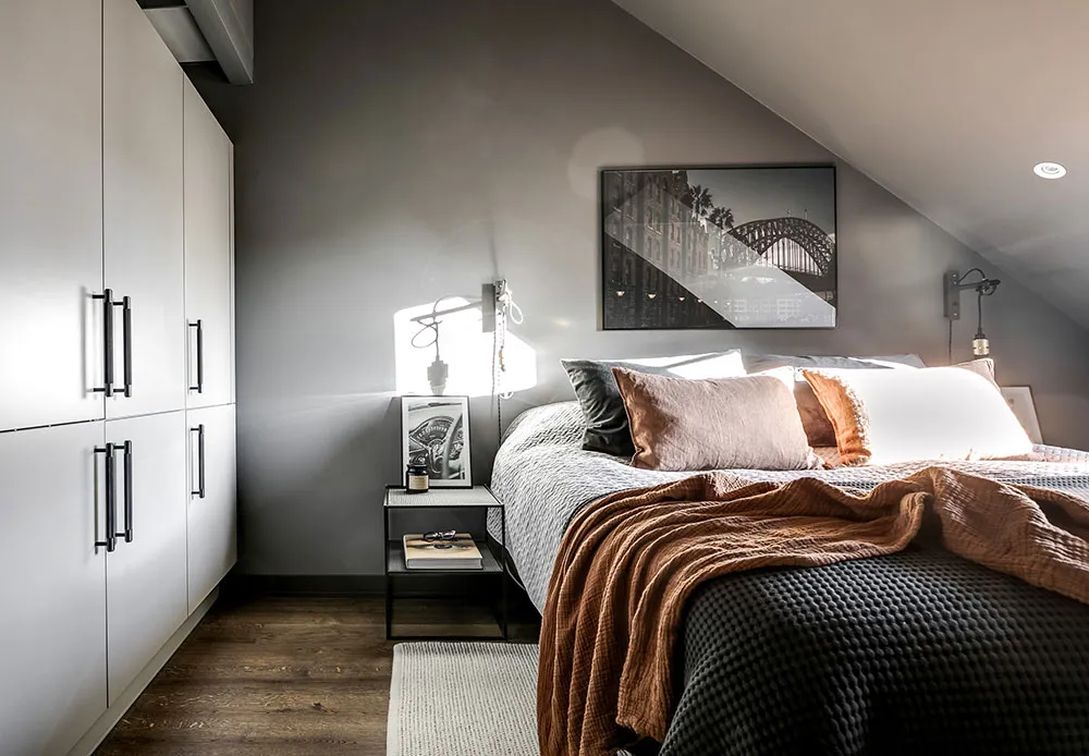 A modern bedroom featuring a bed with a neutral-colored comforter, pillows, and blankets, with a minimalist design and grey w