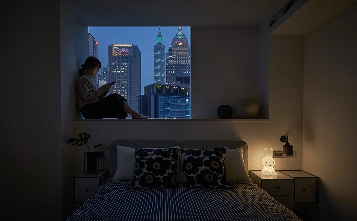 A modern bedroom with textile bedding and a glass window overlooking a city skyline.