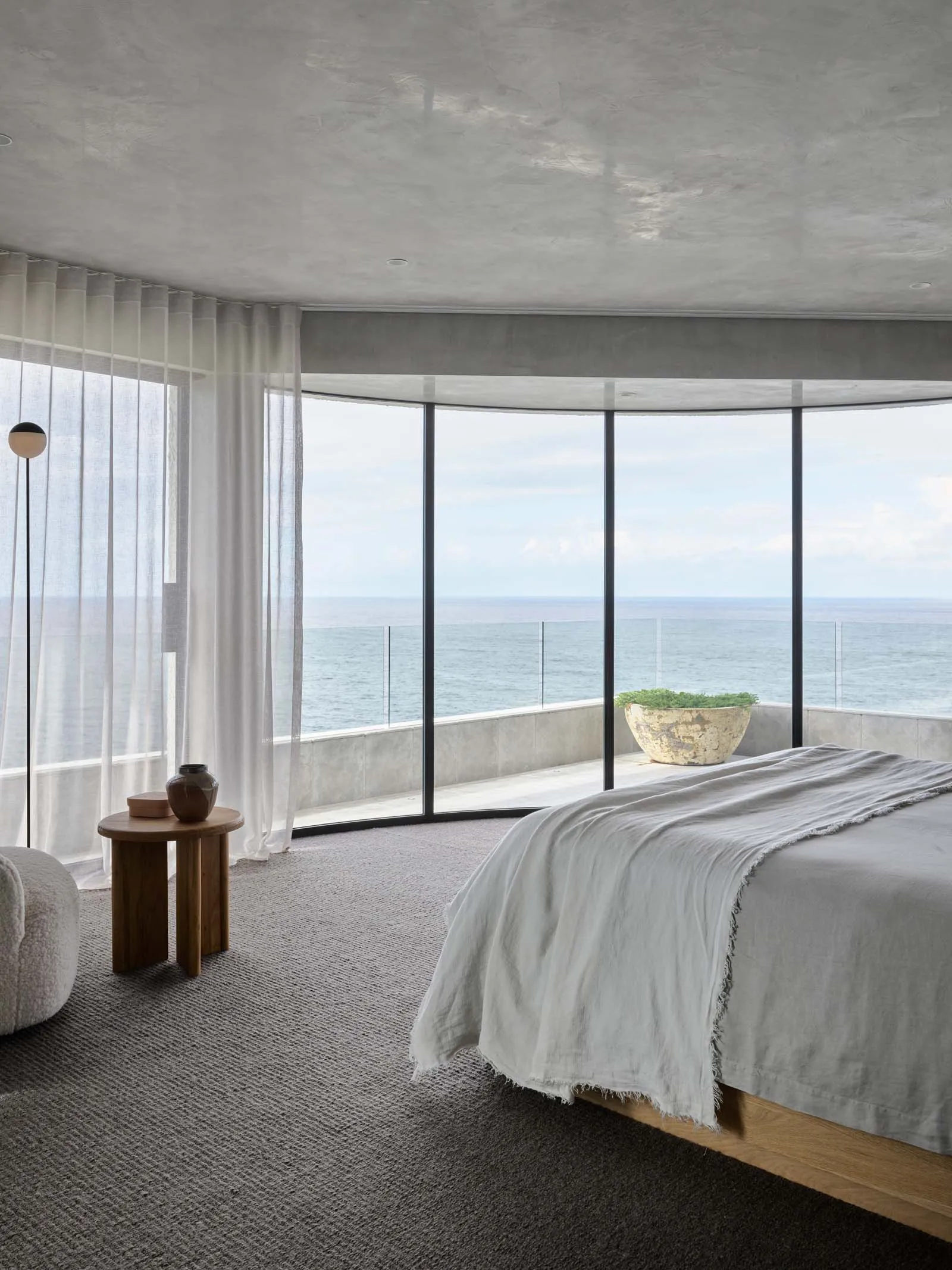 A modern minimalist bedroom featuring a bed in the foreground, with a large window overlooking the ocean, and light-colored c