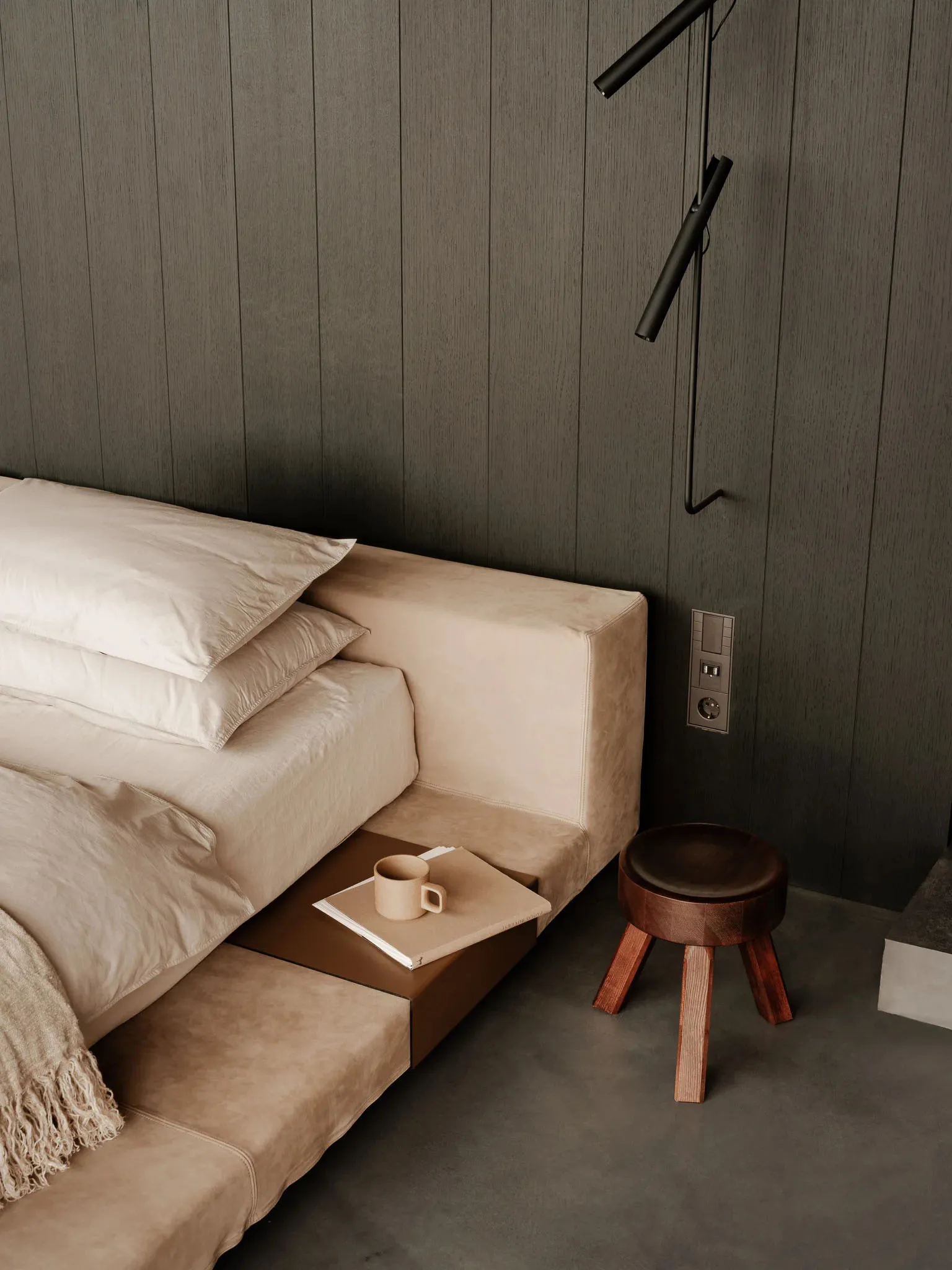 A minimalist bedroom featuring neutral-toned furniture, a modern side table with a coffee mug and book, and a sleek, dark-col