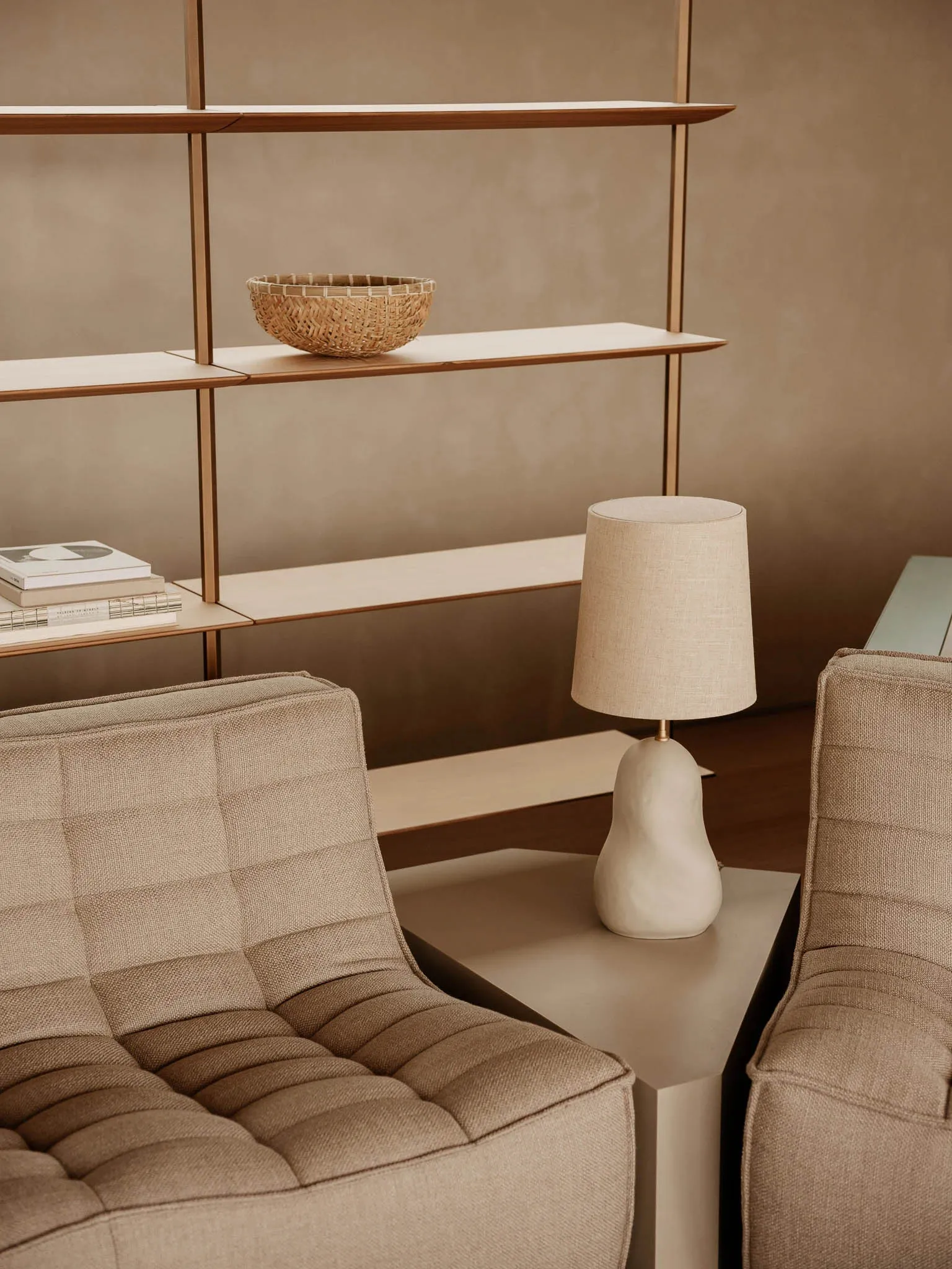 A minimalist living room featuring beige couches and a table lamp with a beige shade, with a beige bookshelf in the backgroun