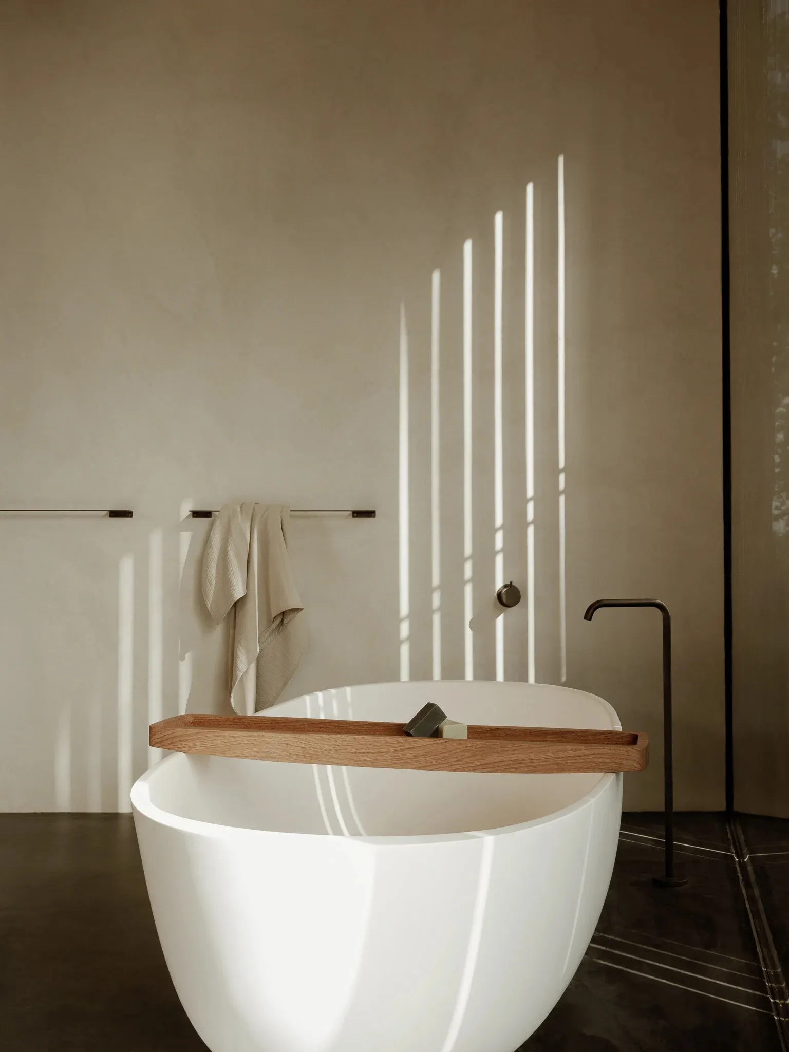A minimalist white concrete bathroom with a freestanding bathtub, a wooden tray, and a towel rack.