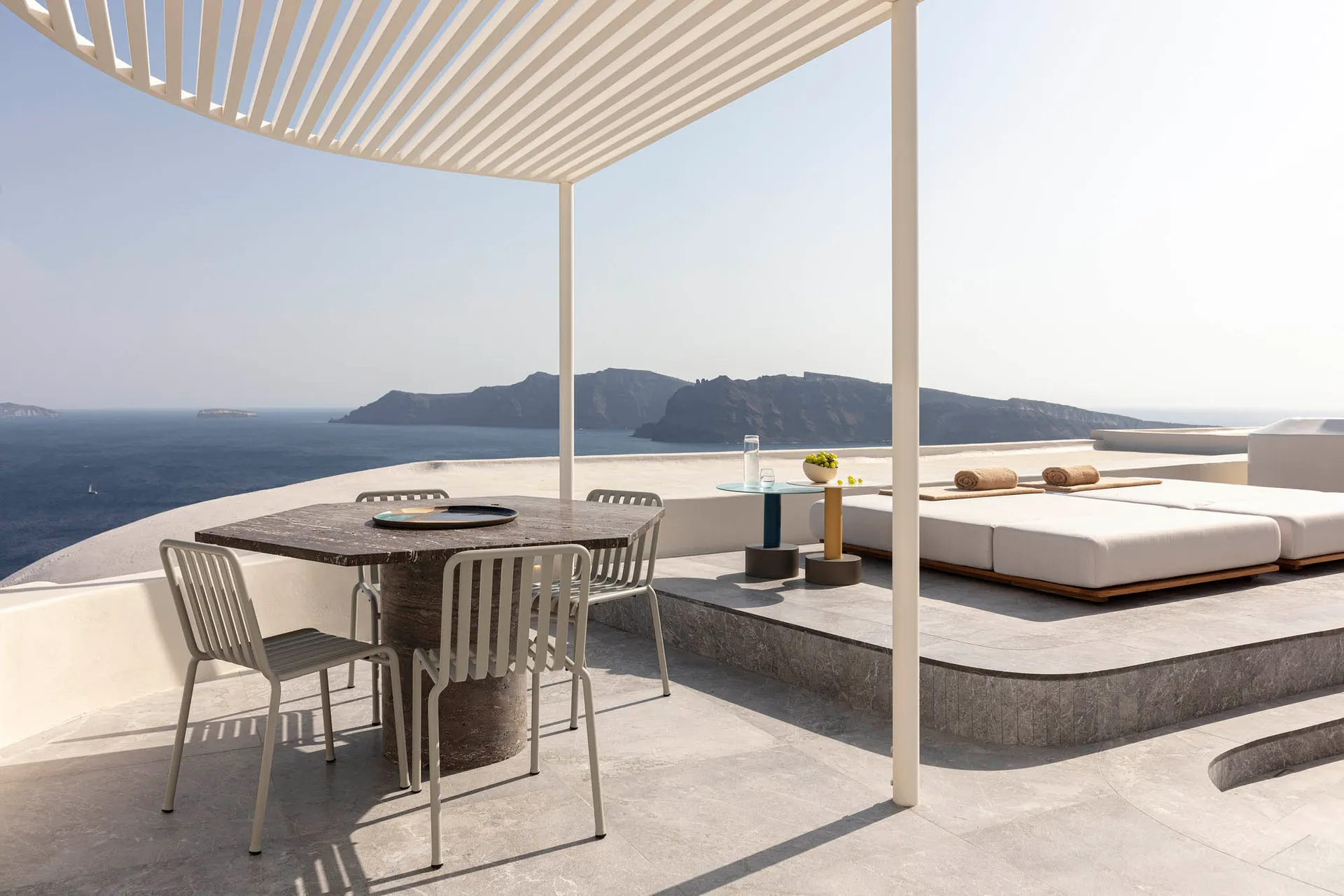 A minimalist residential patio featuring a cement table, metal chairs, glass plant holder, and metal pergola with a view of t