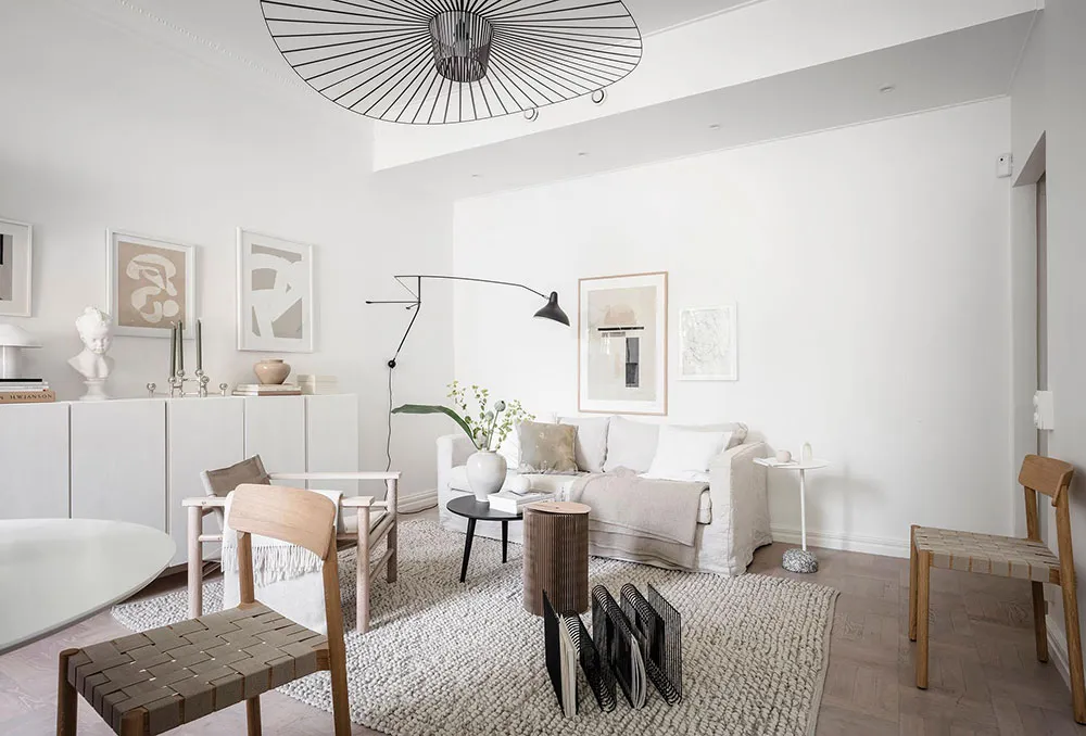 A modern minimalist living room with neutral-toned furniture and decor, featuring a white sofa, wooden chairs, and a black an