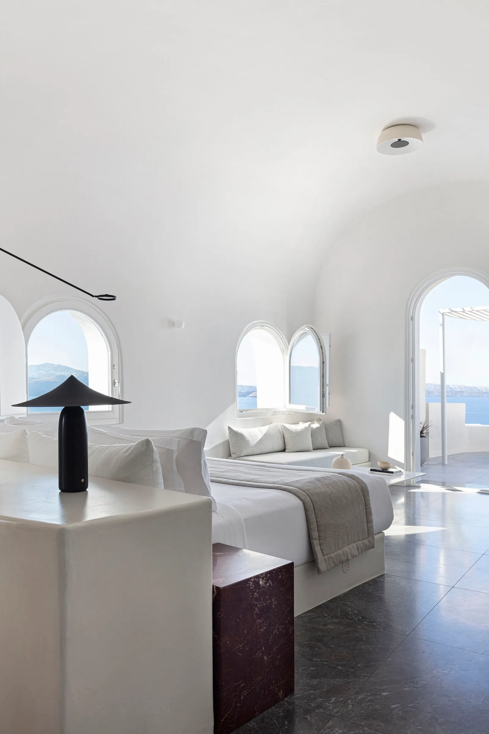 Modern, minimalist bedroom with white walls and concrete floor, featuring a cozy bed and seating area with arched windows.