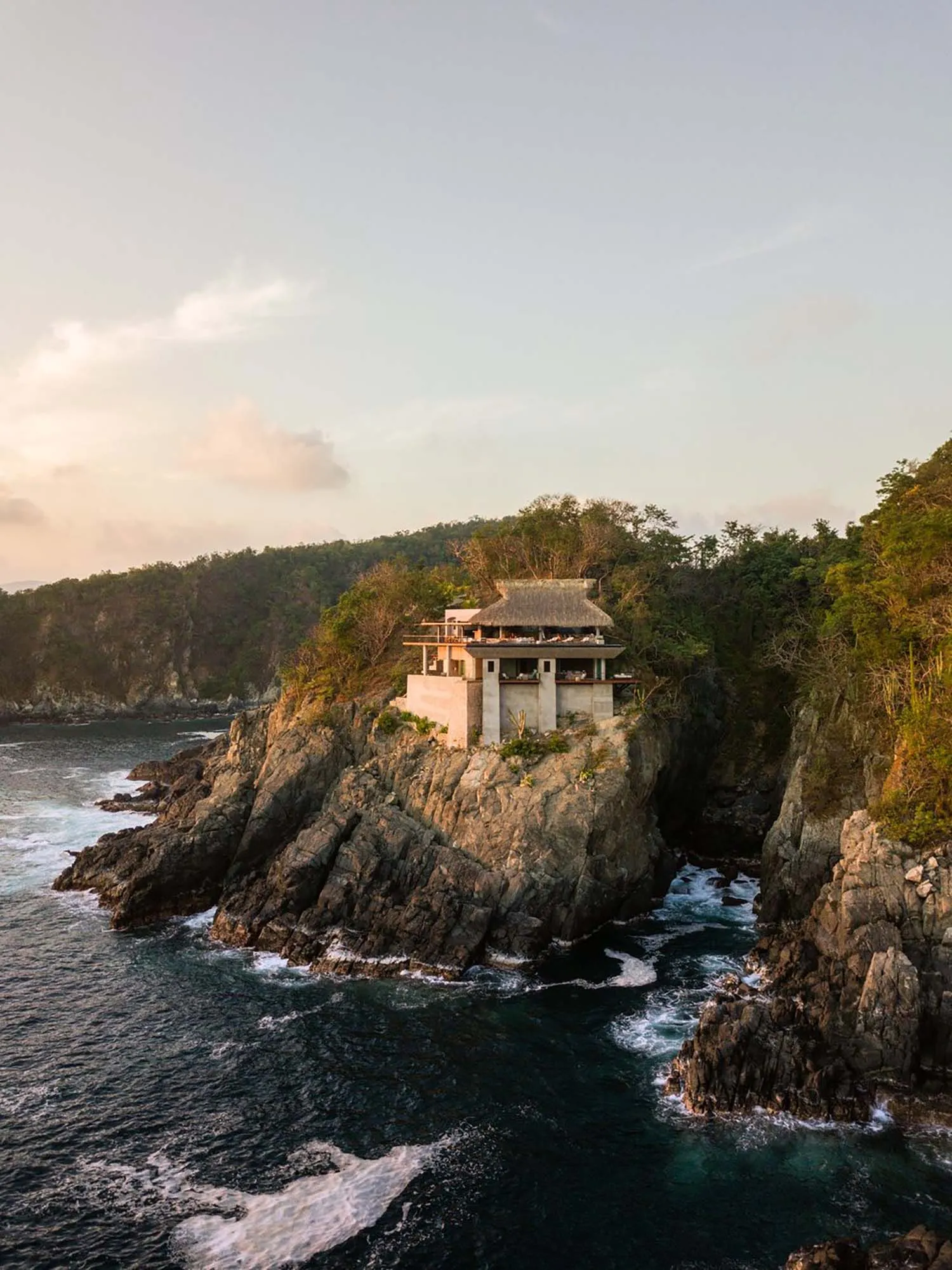 A modern, minimalist cliffside house with a thatched-roof terrace overlooking the ocean.