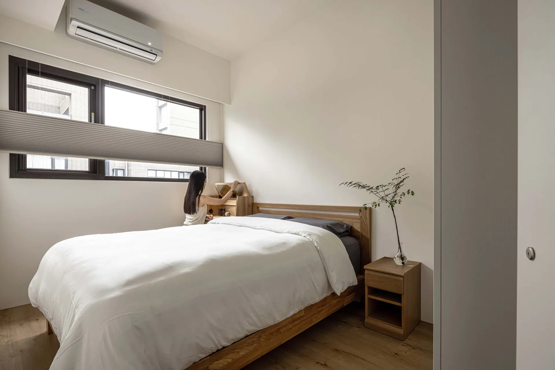 A minimalist bedroom featuring a wooden bed and furniture, with a woman organizing items on the bed.