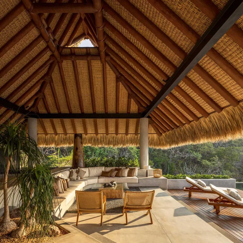 Modern outdoor lounge featuring curved sofas, wicker chairs, and a thatched roof, surrounded by lush greenery.