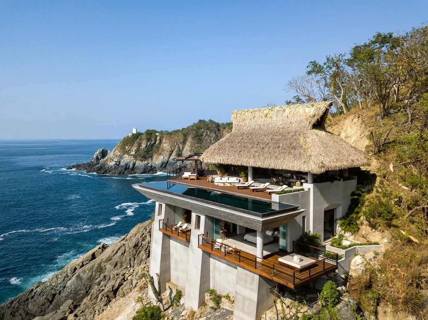 Modern cliffside house with a thatched roof and infinity pool overlooking the ocean.