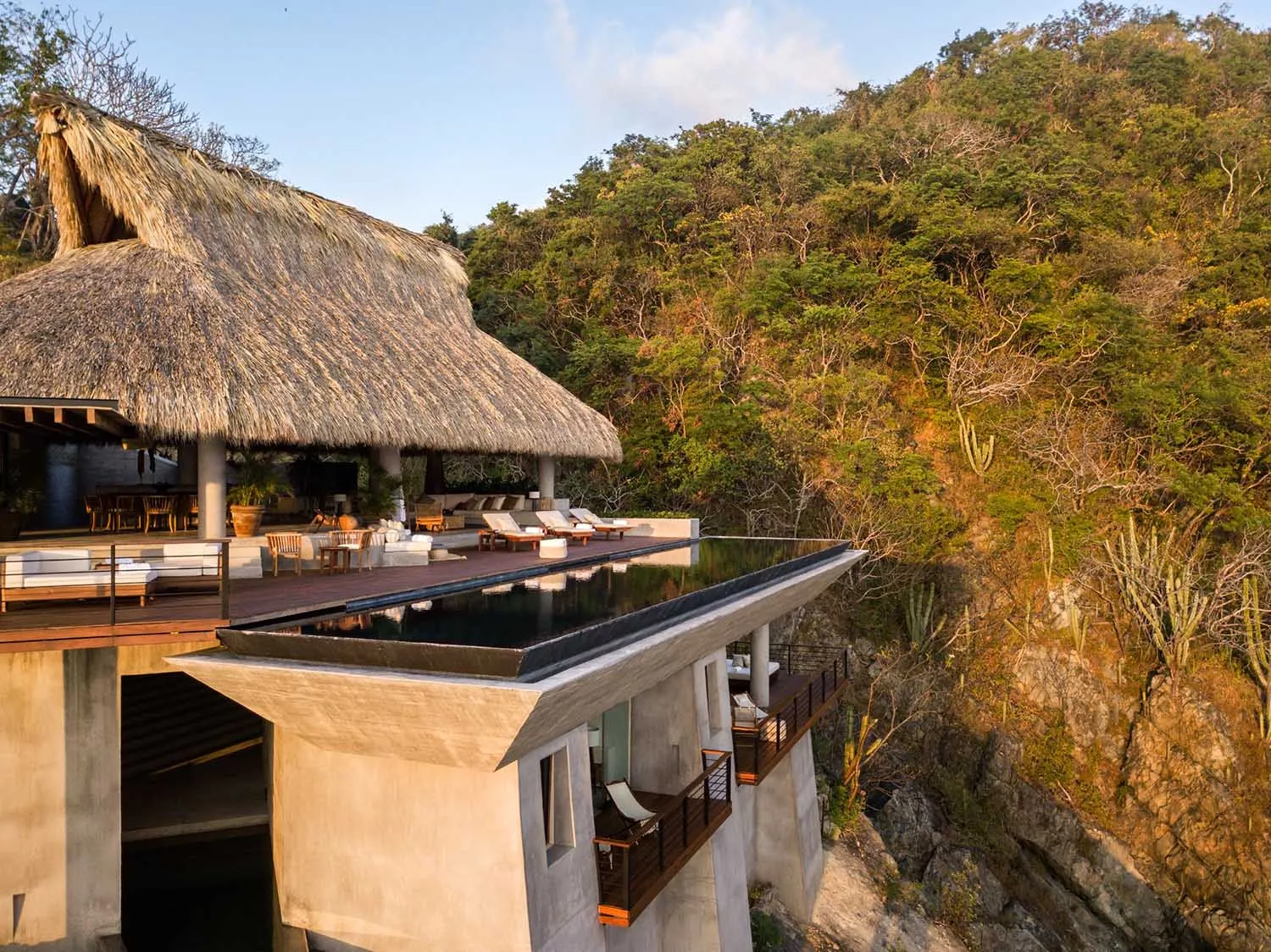 Modern residential exterior with concrete walls, thatched roof, and infinity pool overlooking a forested cliff.