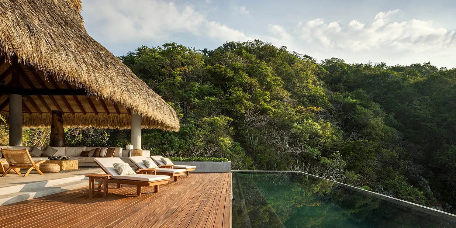 A luxurious outdoor living area with a thatched roof, wooden deck, pool, and lush green surroundings.