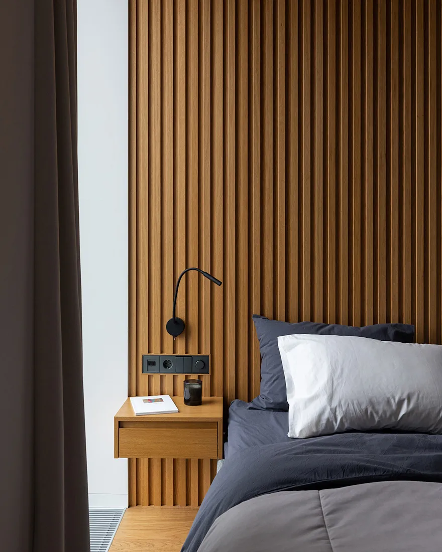 A minimalist bedroom with wooden paneling, a grey bedspread, and a small wooden bedside table with a book and a glass of wate