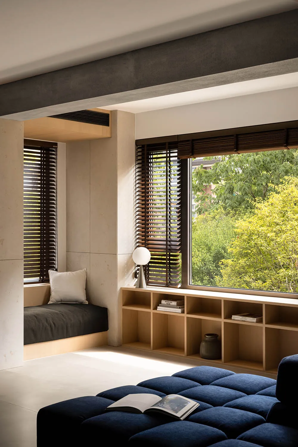 A modern, minimalist living room featuring a window seat with a cushion and a small wooden shelf with books and a plant.