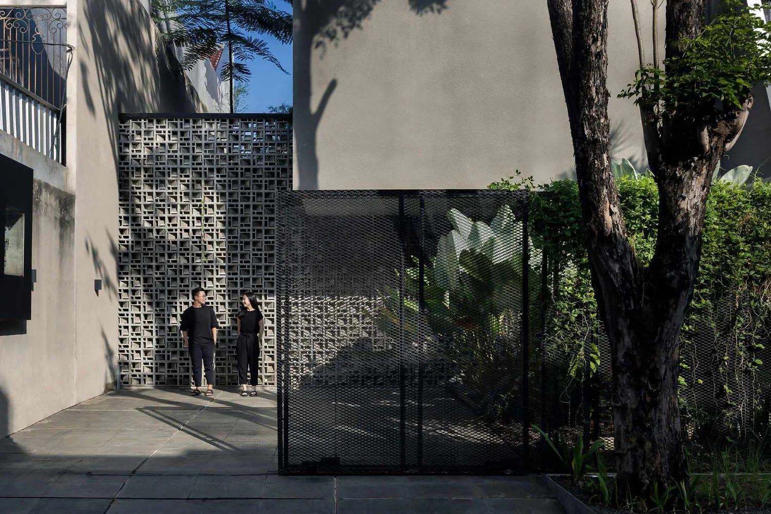 A modern courtyard with concrete walls, a black mesh fence, and lush greenery, featuring two people standing.