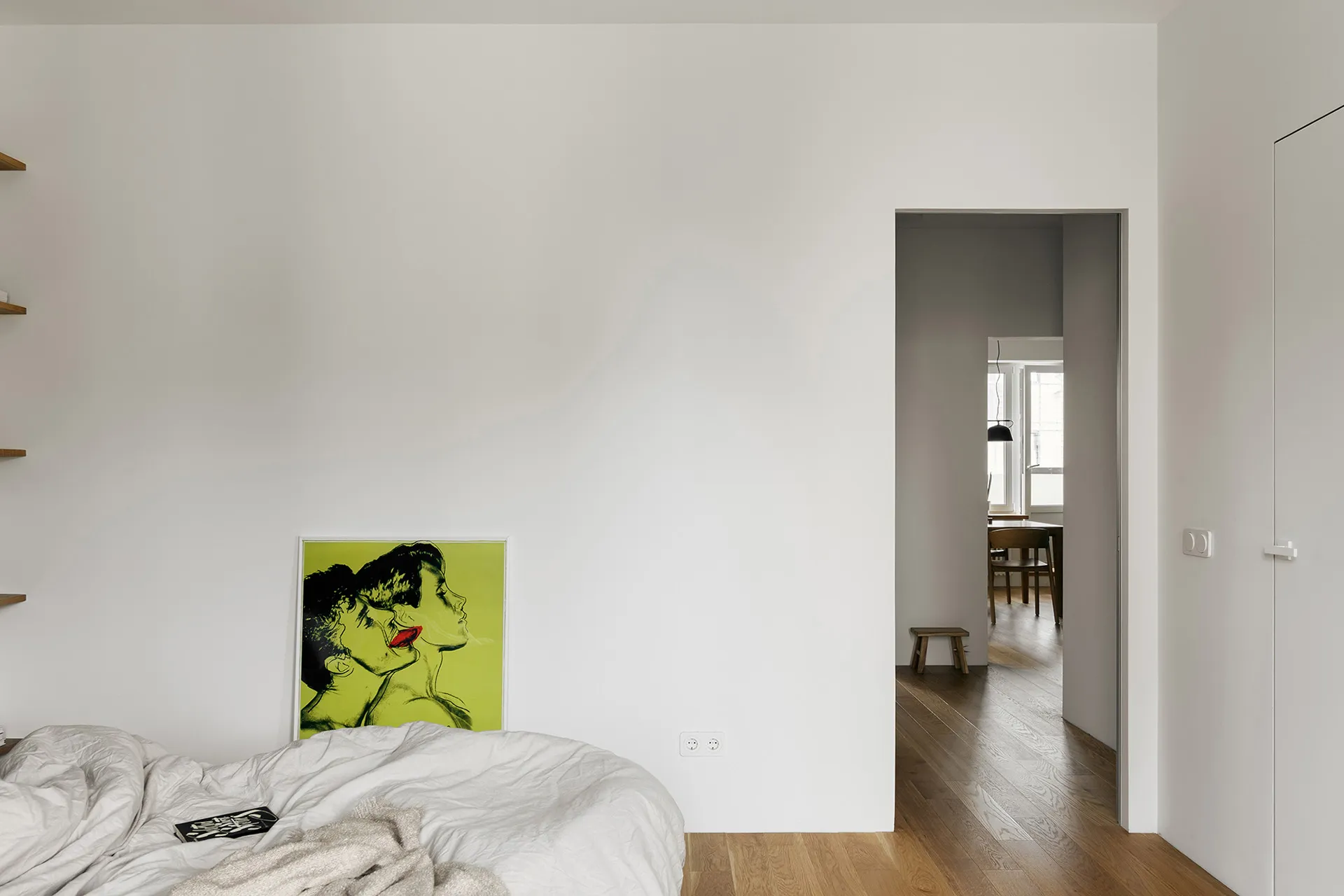 An image of an empty minimalist bedroom with white walls, wood flooring, and a green and yellow framed artwork.