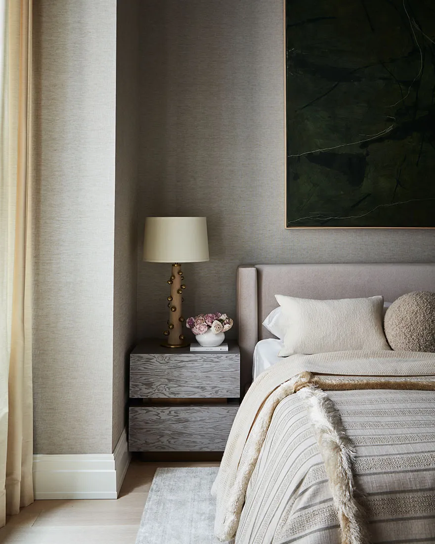 Modern bedroom with neutral textiles and a wooden nightstand with a table lamp and flowers.