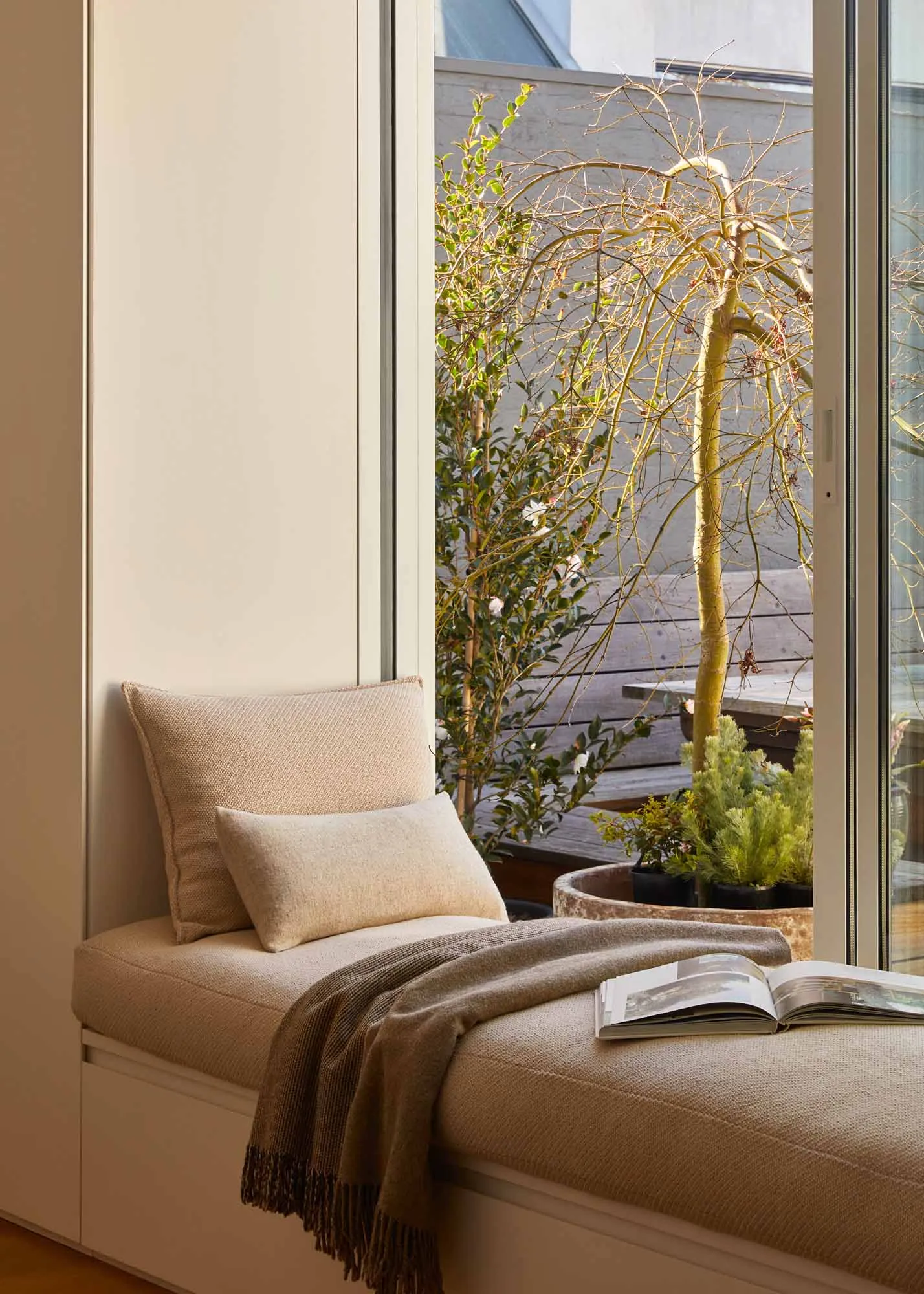 A modern bedroom with a beige sofa adorned with cushions, placed by a window overlooking an outdoor area with plants.