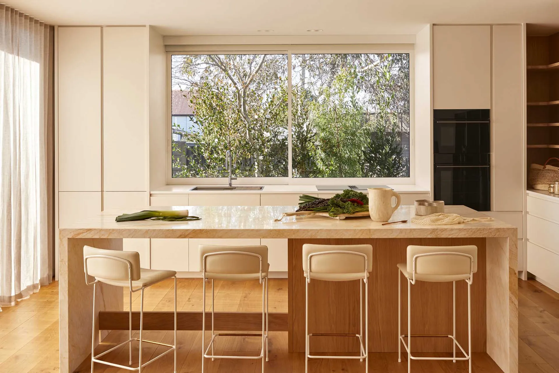 A modern kitchen with a marble countertop, stools, a kitchen island, and a large window overlooking greenery.