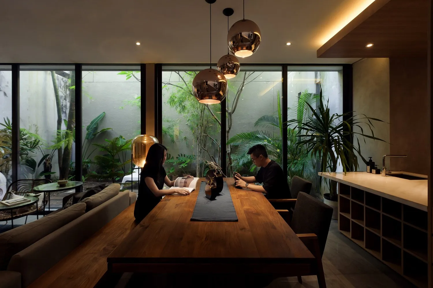 A modern living room with a wooden table, glass pendant lights, and green plants.