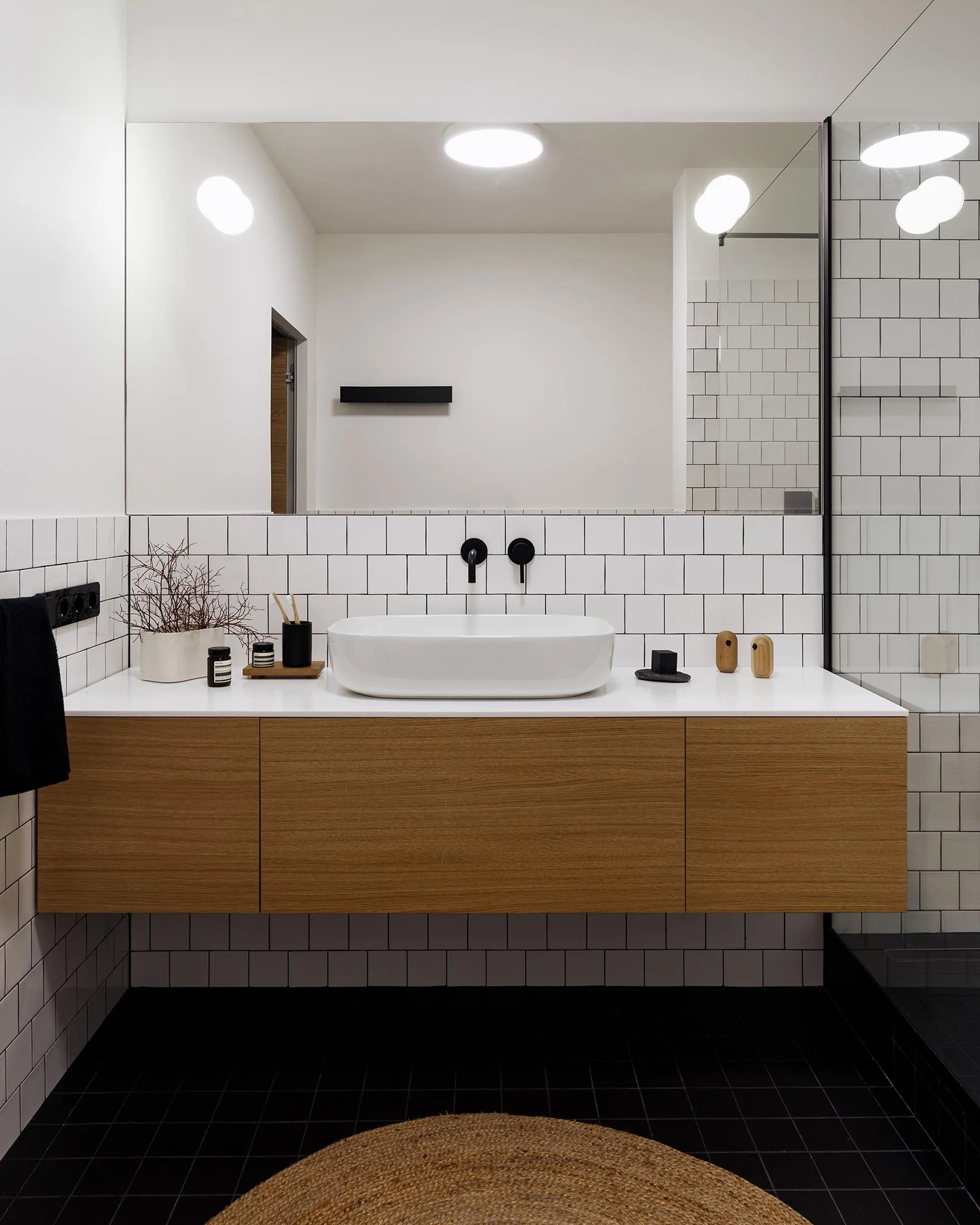 A modern minimalist bathroom with white tiles, wooden cabinets, and a wooden vanity with a white sink. The floor is covered w