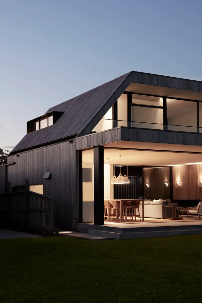 A modern house with black siding and large glass windows illuminated against a twilight sky.