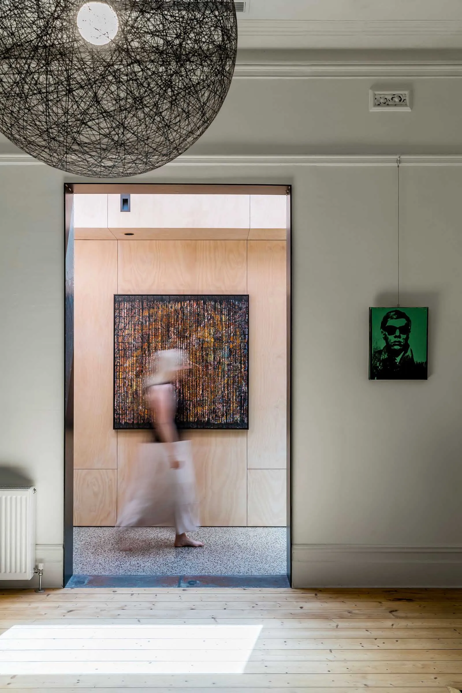 A blurred figure walks through a modern entryway with a wood-paneled wall and abstract painting.