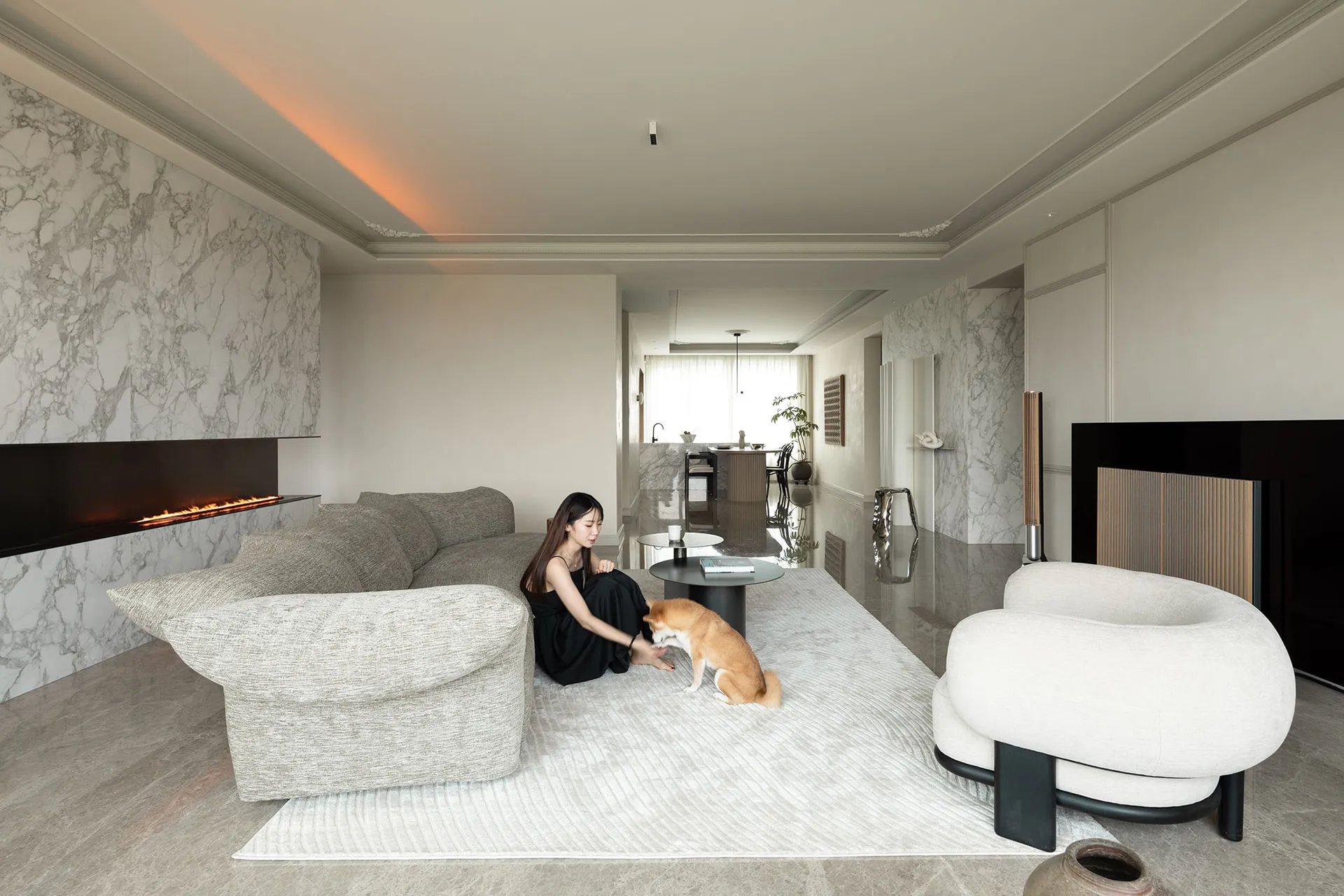 A modern living room with a marble accent wall, grey L-shaped sofa, and a woman sitting with a dog on a plush rug.