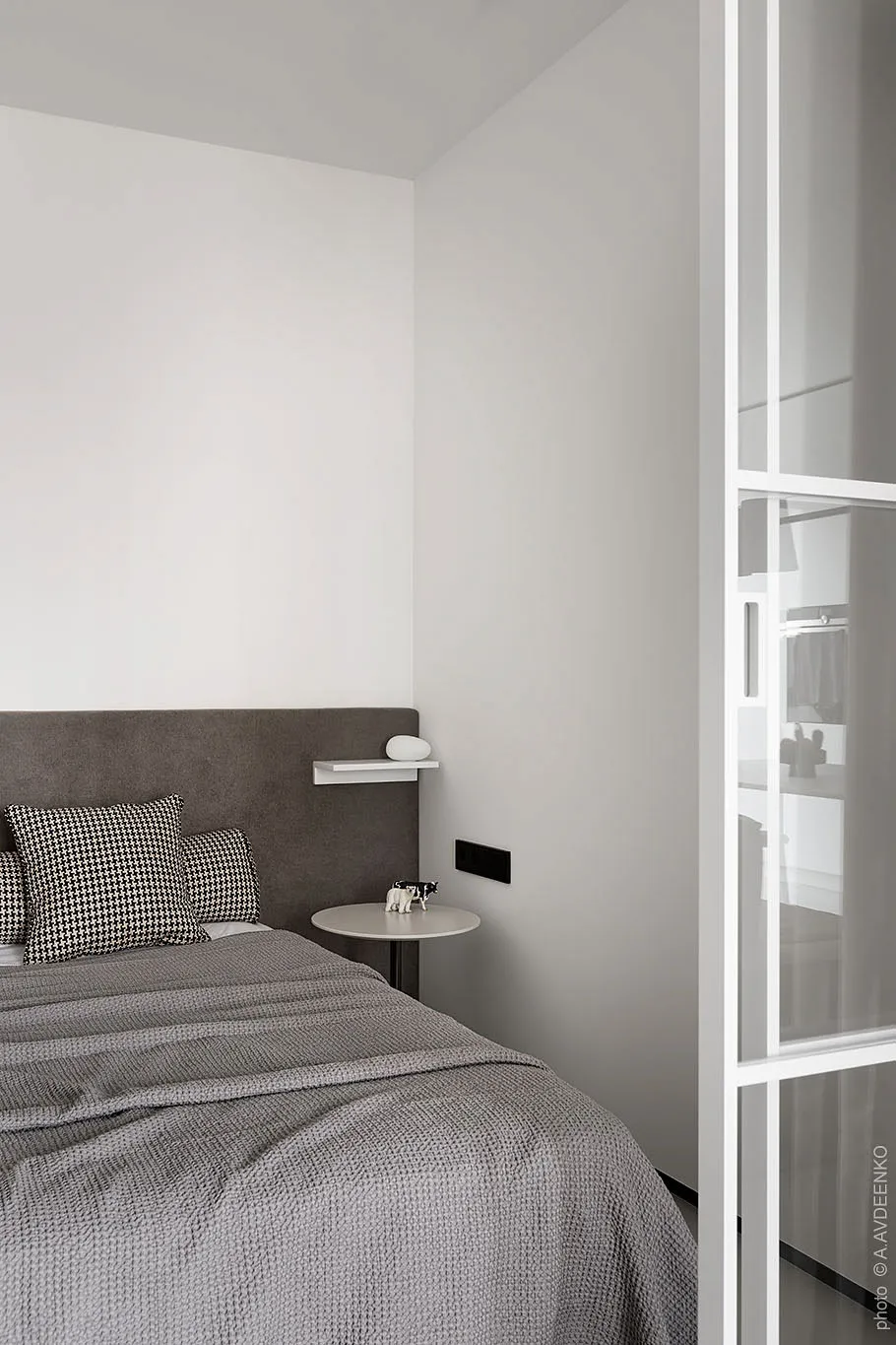 Modern minimalist bedroom with white walls, a grey fabric bed, and textured bedding, featuring a glass closet door.