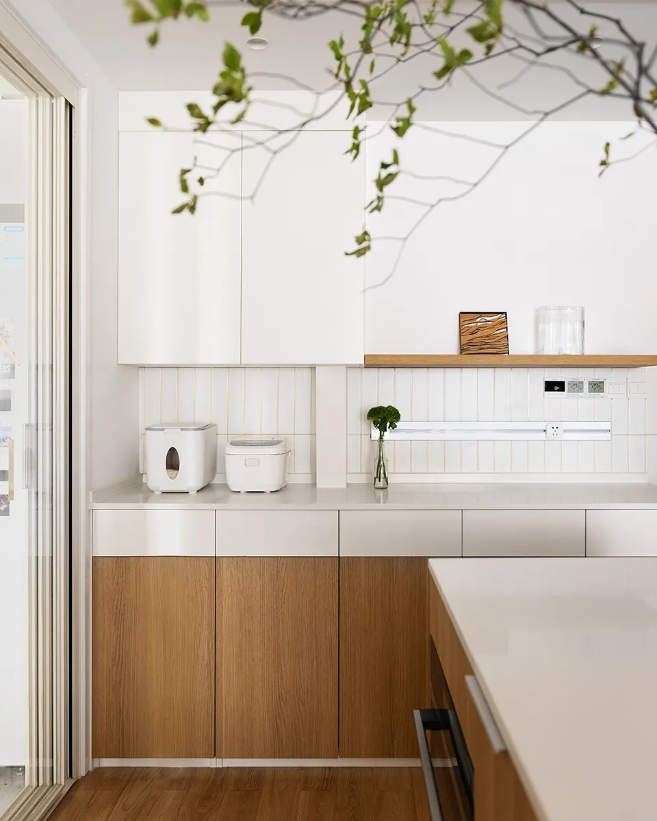 A modern, minimalist kitchen featuring sleek, white cabinetry and wood accents, with a white countertop and a window providin
