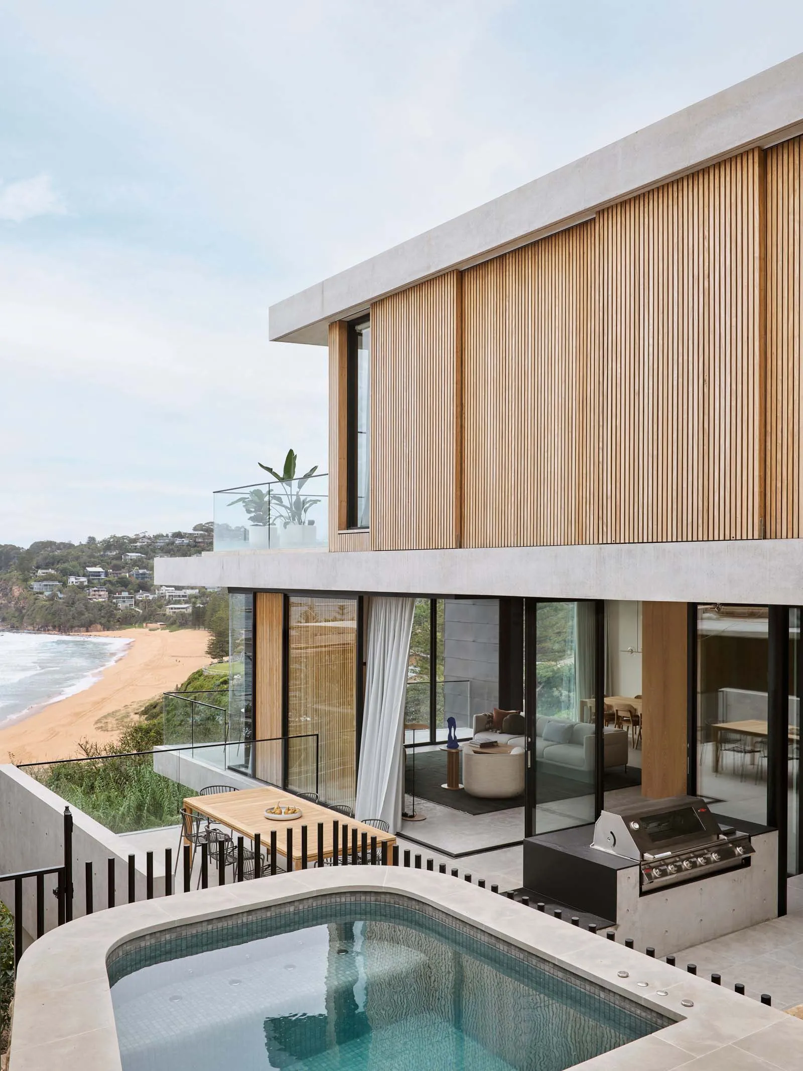 Modern residential exterior with glass walls, wooden slats, and a swimming pool overlooking a beach.