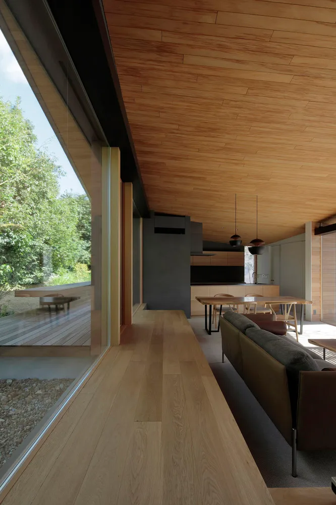 Modern living room with wooden walls and flooring, large windows overlooking greenery, contemporary furniture, and a minimali
