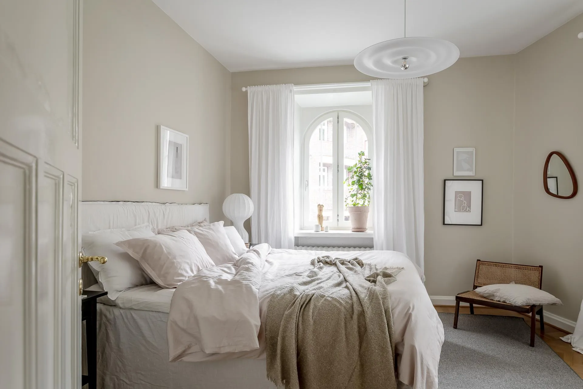 A minimalist bedroom with fabric bedding and wooden furniture, featuring an arched window and a plant on the windowsill.