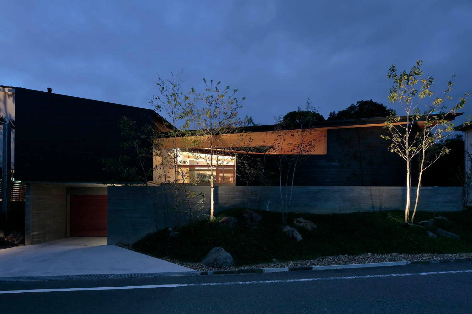 A modern house with a black exterior made of concrete and wood, featuring a driveway and tree lighting.