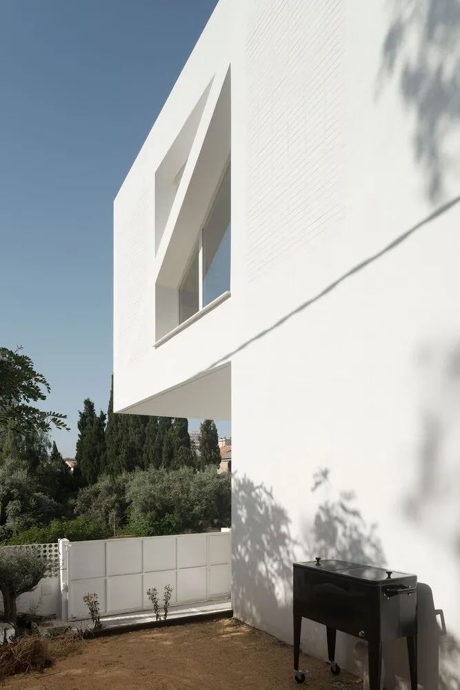 A modern house with a white exterior made of concrete, featuring a unique window design and a black barbeque grill in the for