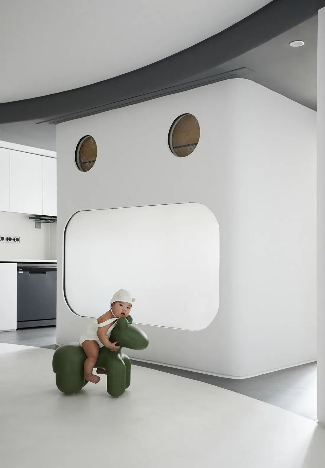 A child sits on a green, plastic toy horse in a minimalist play area with white walls and floor.