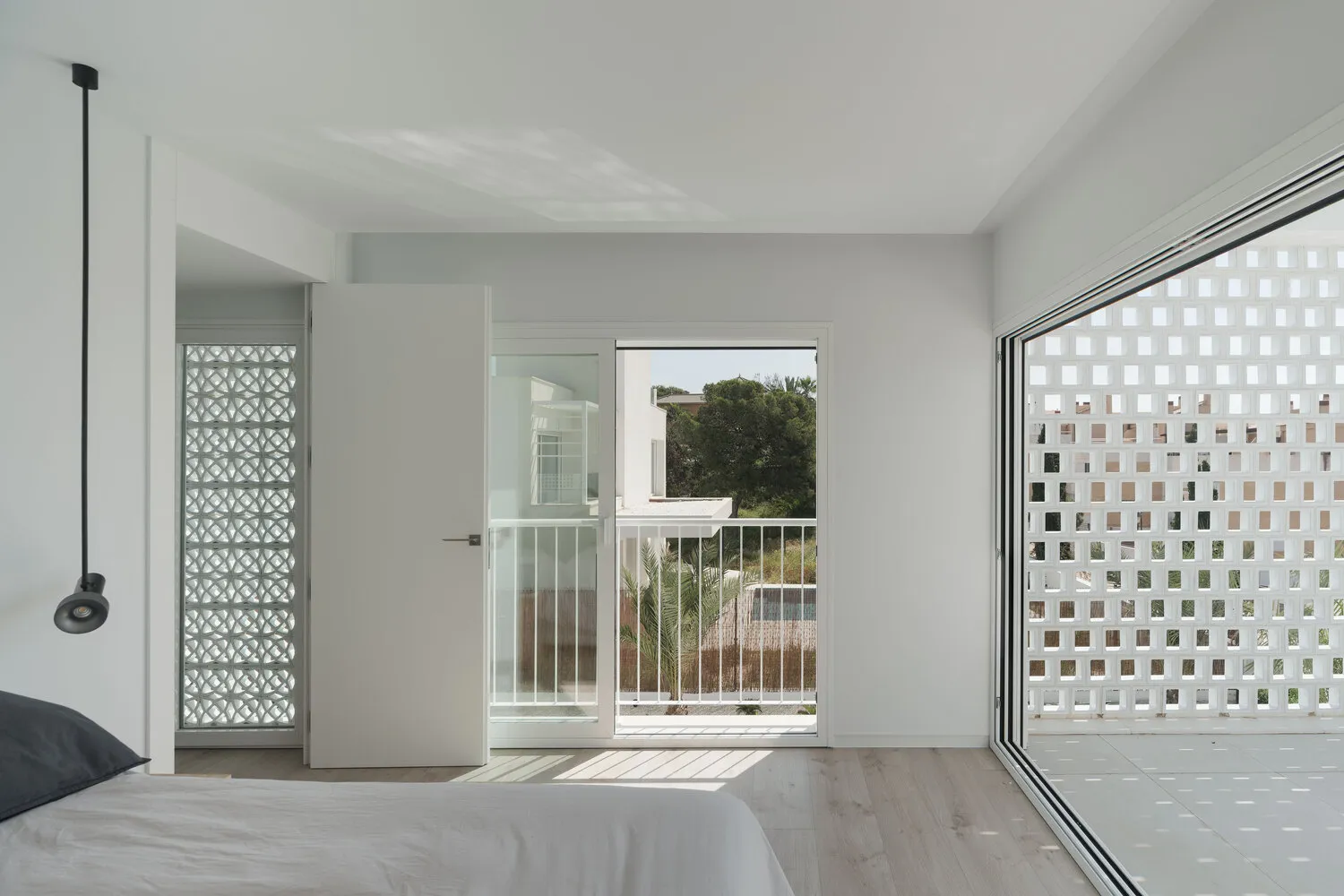 A minimalist bedroom with white walls, wood-laminate flooring, and a large window overlooking a balcony with a metal railing.