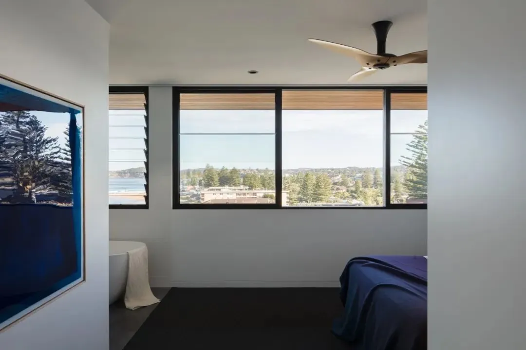 Modern minimalist bedroom featuring a large window with a view of the ocean and trees, a blue bedspread, and a large framed p