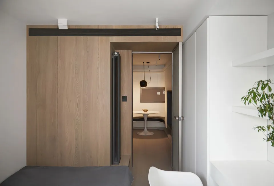 A minimalist bedroom with white walls, a grey bed, and a white chair, featuring a walnut wall panel and a white floating shel