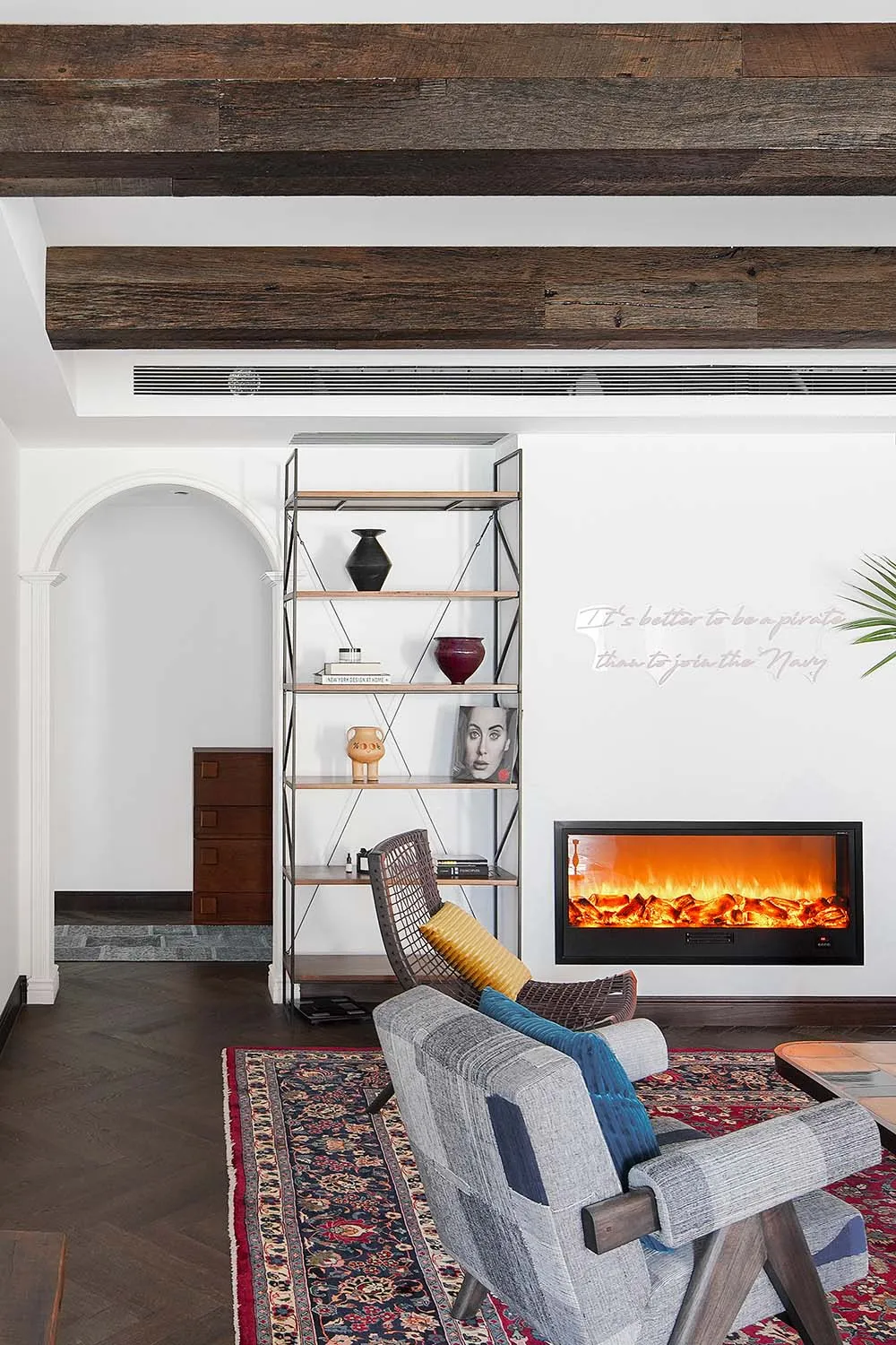 A minimalist living room featuring a wooden fireplace, bookshelf, and comfortable seating area on a patterned rug.