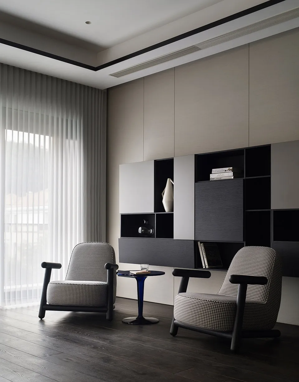 A modern minimalist living room featuring two patterned armchairs, a wooden floor, and a large window with sheer curtains.