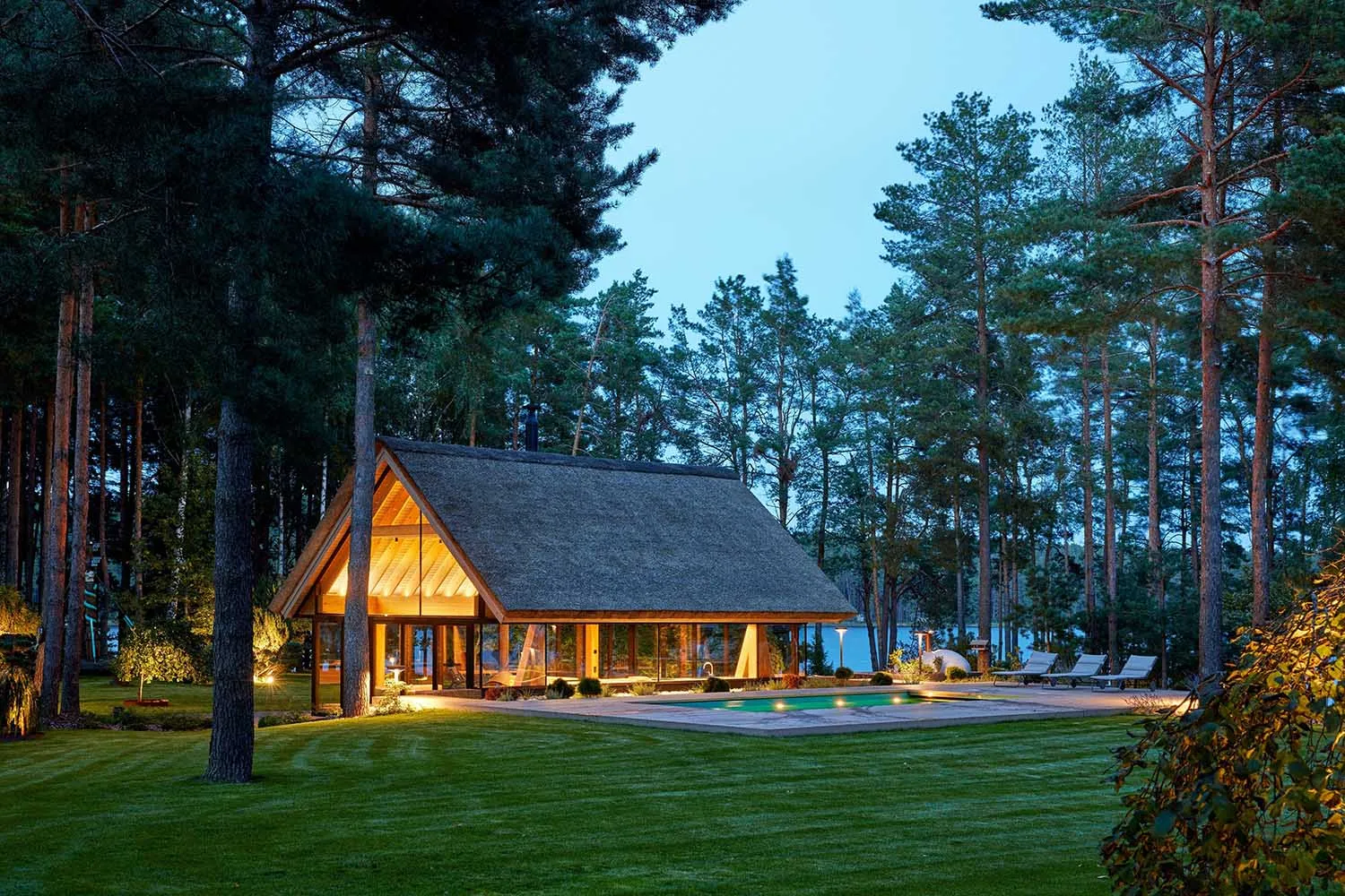 A modern country house with a thatched roof and wooden exterior, surrounded by tall trees and featuring a pool.