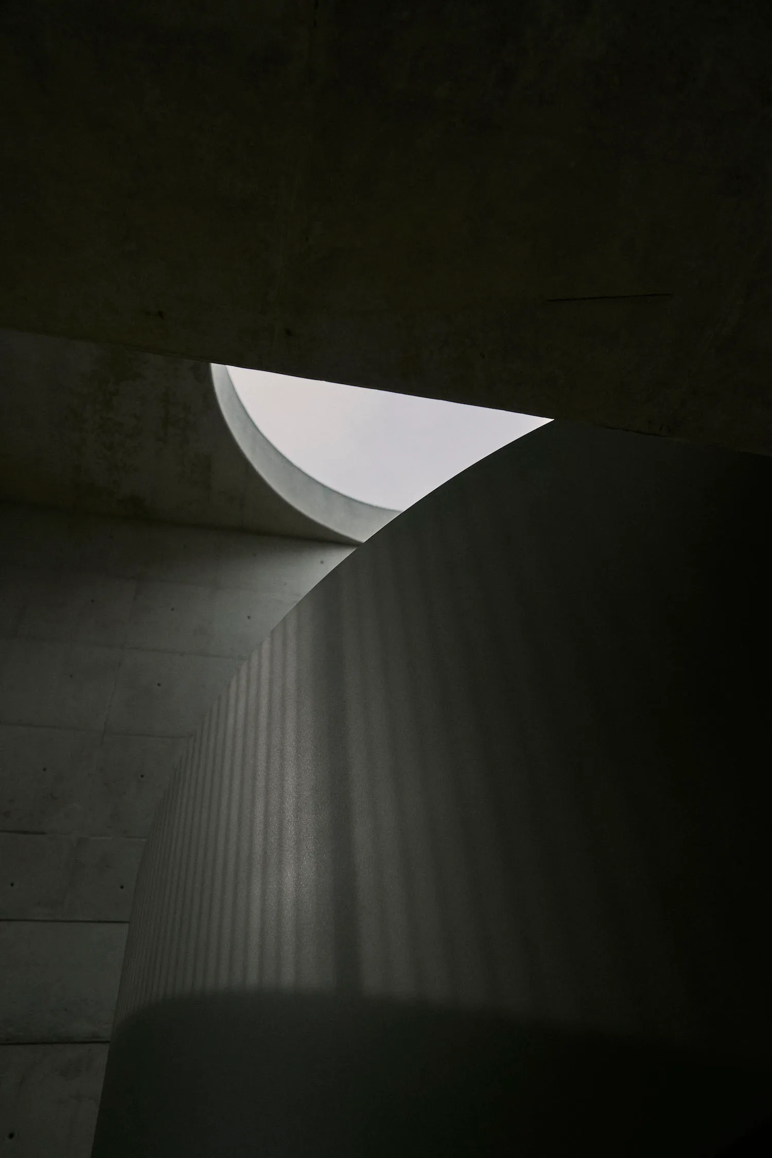 Circular concrete structure with light filtering through, casting vertical shadows on the wall.
